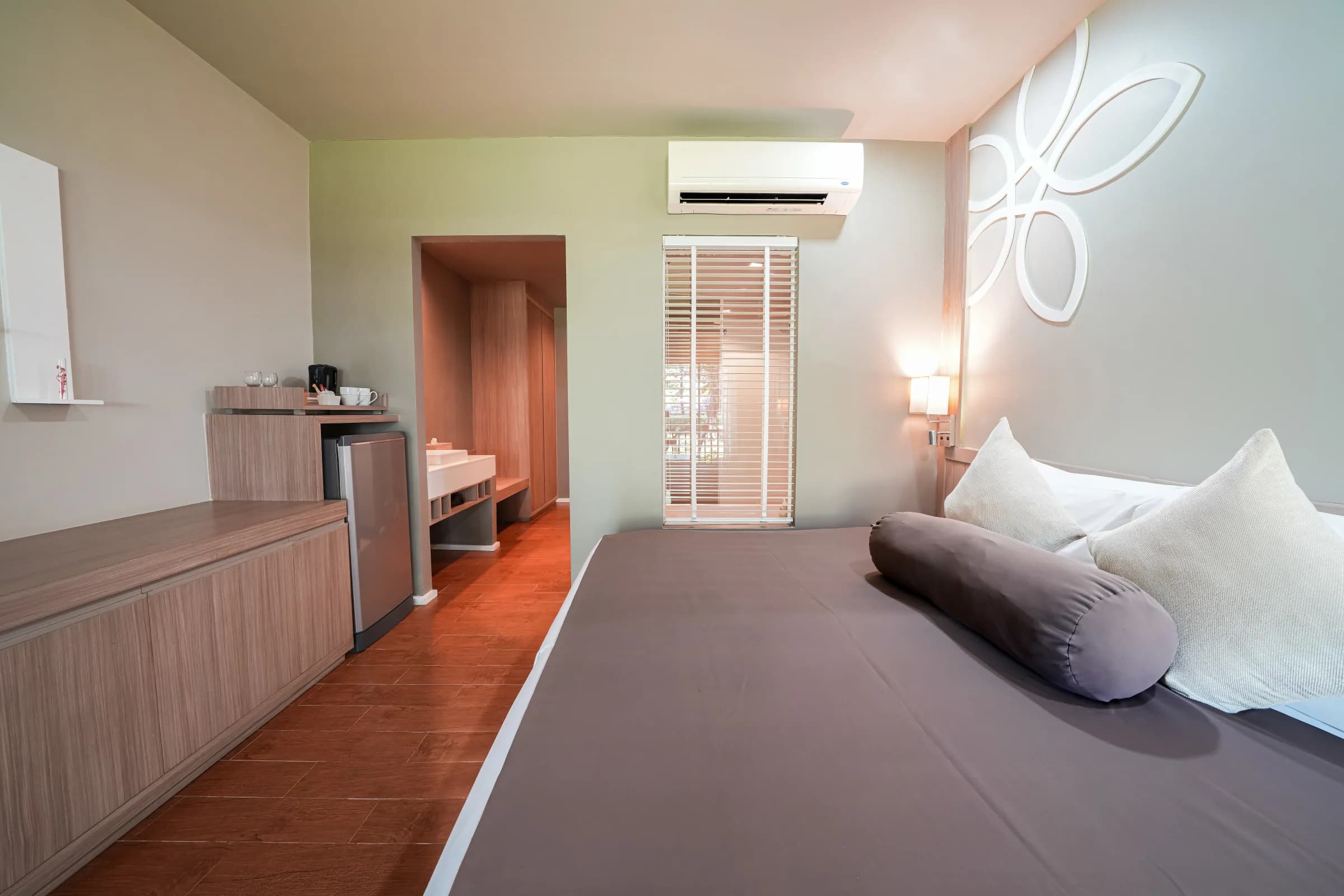 Hotelzimmer mit grauem Bett, weißen Kissen und einer Holzkonsole mit Minibar. Blick auf ein offenes Badezimmer.